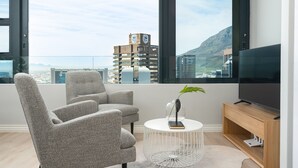 Un salon moderne doté d’une grande fenêtre, de fauteuils gris à revêtement en tissu, d’une table basse ronde, d’un meuble pour la télévision et offrant une vue sur un paysage urbain et des montagnes.