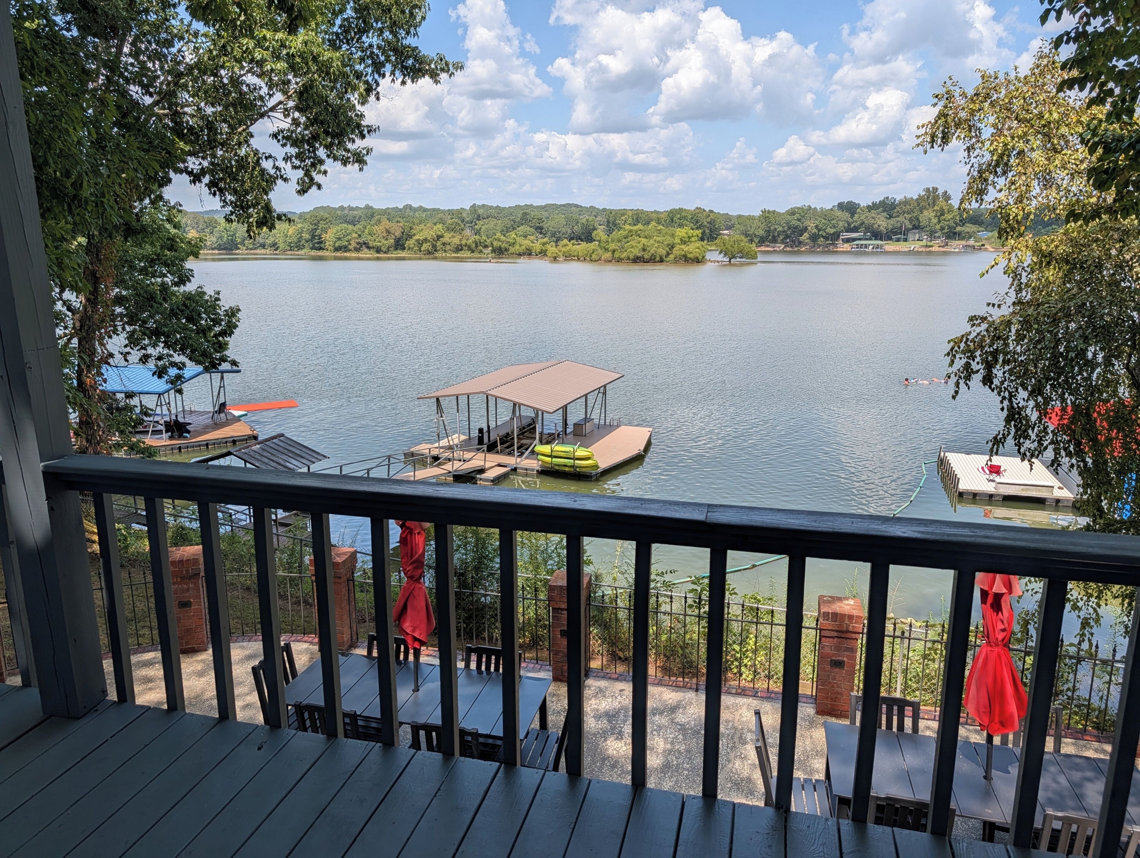 A lakeside deck with tables and chairs, a covered dock, and a view of the lake and distant trees.