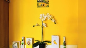 A shelf with greeting cards, a potted orchid, and a small painting on a yellow wall.