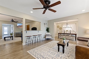 A spacious living room with a wooden floor, a patterned rug, a ceiling fan, a dining area with a chandelier, and a kitchen in the background.