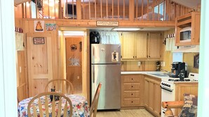 A cozy kitchen with wooden cabinets, a stainless steel refrigerator, and a dining area with a table and chairs. - Assoc beach, Lakes, Naples Causeway, restaurants, shops, boat- jet ski rentals. (Naples)