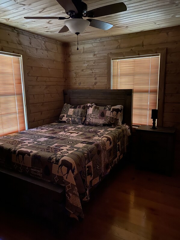 A bedroom with a wooden ceiling, a ceiling fan, a bed with a patterned quilt, and two windows with blinds.