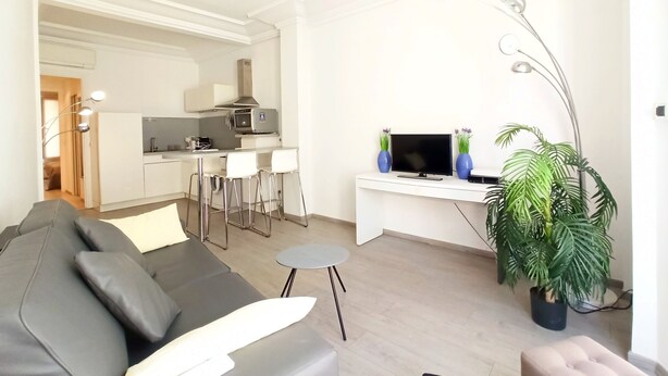 Ein modernes Wohnzimmer mit grauem Sofa, einem kleinen runden Tisch und einem Fernseher auf einem weißen Schreibtisch. Im Hintergrund ist eine Küche mit weißen Schränken und einer Mikrowelle zu sehen.