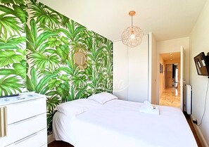 Quarto com cama, penteadeira, espelho, parede com padrão de folha tropical e luminária no teto.