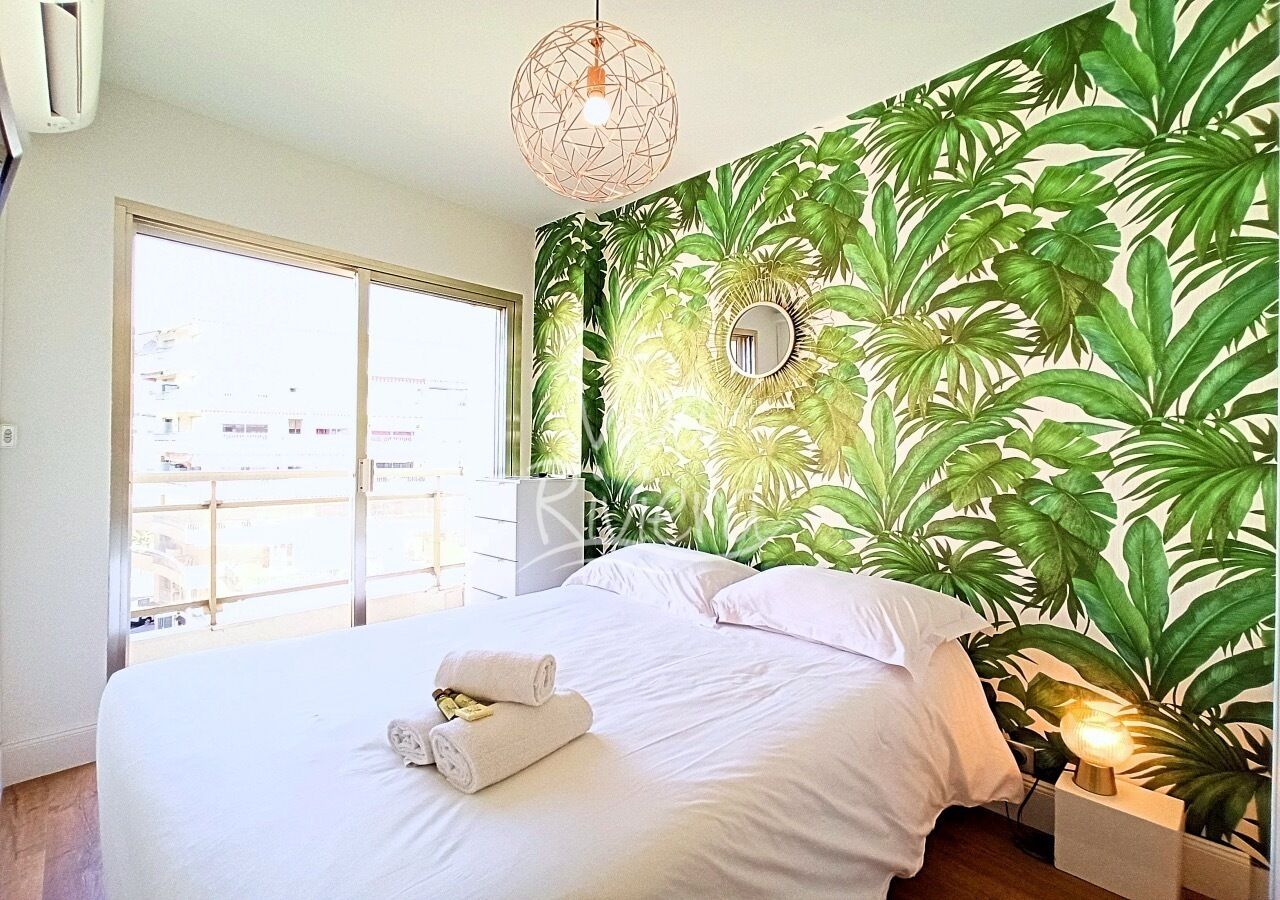 A bedroom with a large bed, a view of the city through a balcony, and a tropical-themed wall mural.