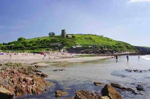 A beach with rocky foreground, a grassy hill with a building, and people in the distance.