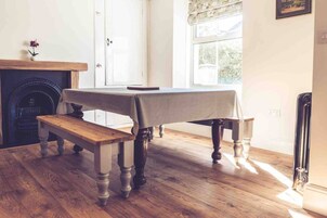 A room with a wooden floor, a table with a tablecloth, a bench, a fireplace, and a window with a view of greenery.