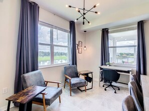 A modern office space with a desk, two chairs, a hanging light fixture, and large windows offering a view of a park.