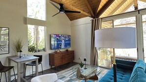 A spacious living room with a high ceiling, wooden beams, a large TV, a sofa, a coffee table, and a dining area with chairs and a table. - Condo with amazing views and 3 pools (Indian Wells)