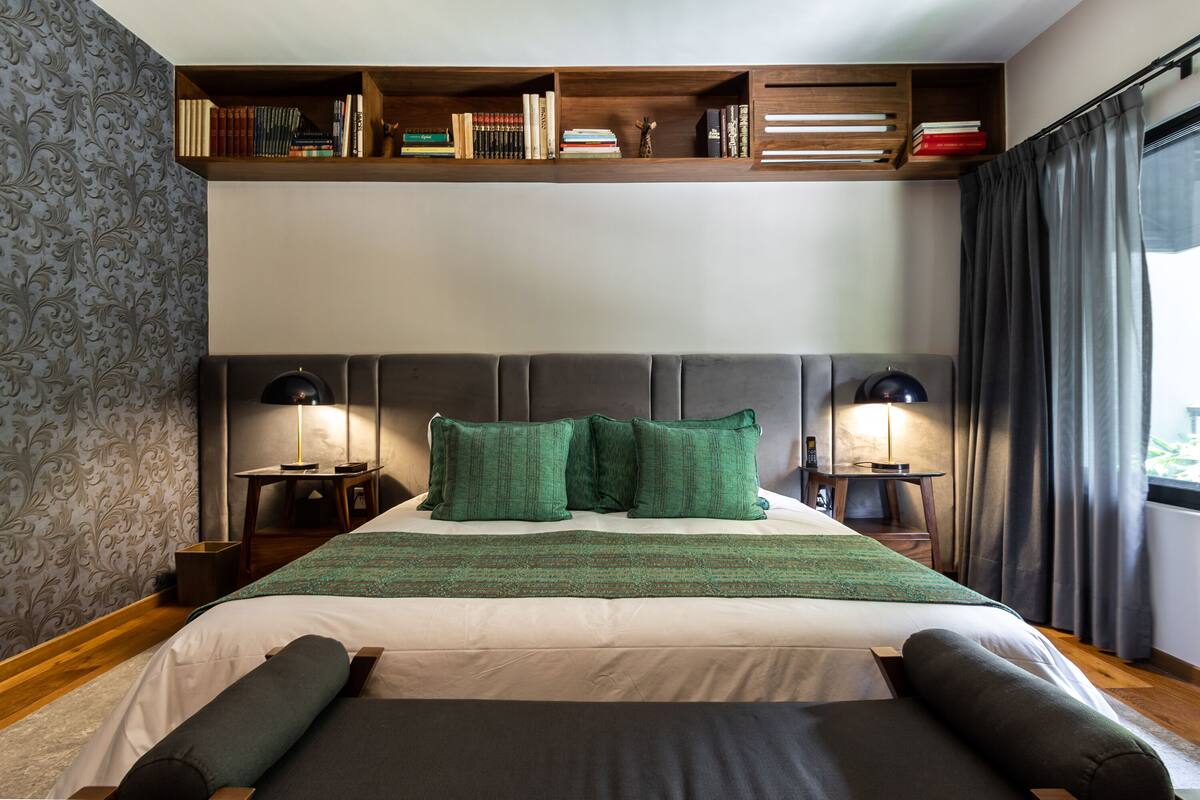 Cama bem arrumada com almofadas verdes e manta listrada, uma estante acima e duas luminárias de cabeceira.