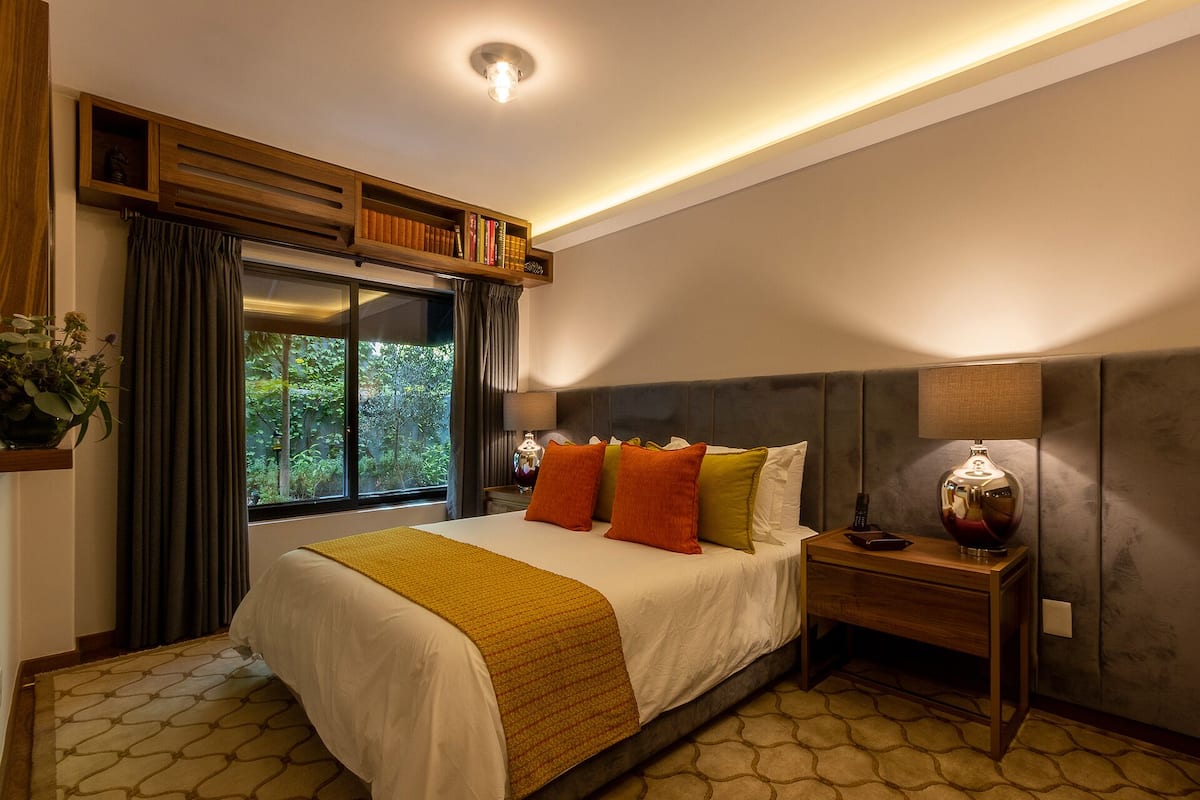 Quarto de hotel moderno com uma cama grande, uma mesa de cabeceira, um abajur e uma janela com cortinas.