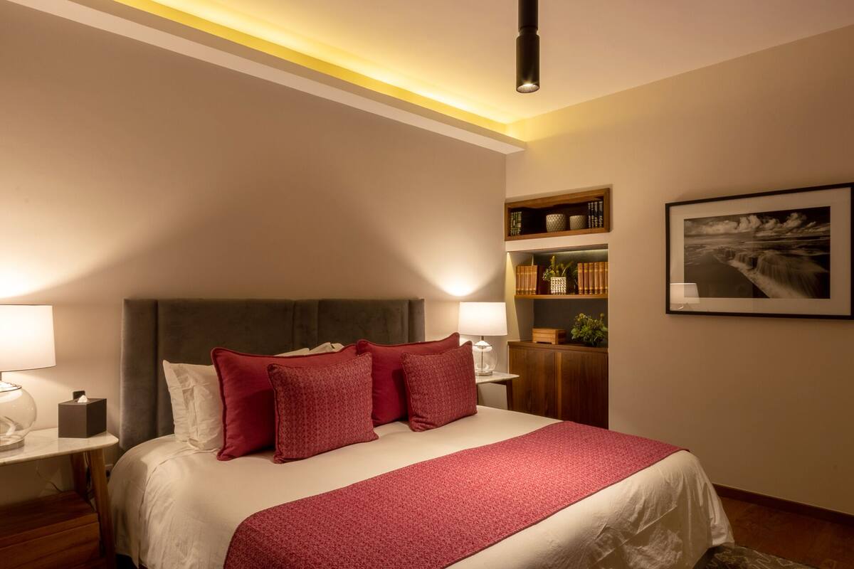 Quarto de hotel com uma cama grande, mesa de cabeceira com abajur, estante de livros e um quadro na parede.
