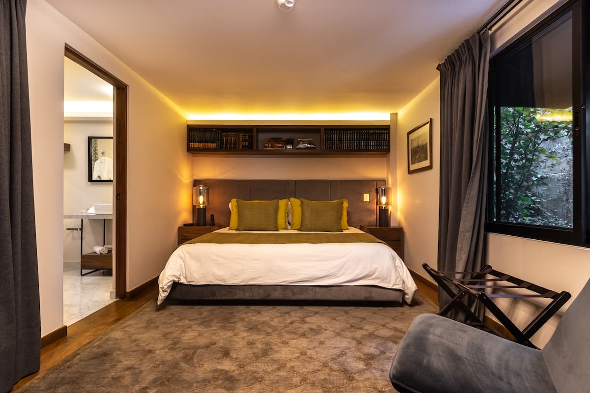 Quarto com cama, mesas de cabeceira, uma cadeira, janela com cortinas e vista para o exterior.