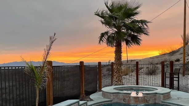A swimming pool with a hot tub, surrounded by a wooden fence, with a sunset view.