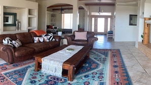 A spacious living room with a brown leather sofa, a wooden coffee table, and a patterned rug. There is a television on a wooden stand to the right.