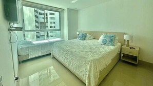 A bedroom with a large bed, a nightstand, a TV mounted on the wall, and a window with a view of a building. - Apartamento Vista al Mar (Cartagena)
