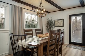 Un coin repas avec une table en bois, des chaises et un lustre. On y trouve des fenêtres avec des rideaux, une porte avec un vitrage et un tableau encadré au mur.