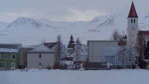 Une ville enneigée, avec un clocher d’église, des maisons et des montagnes en arrière-plan.
