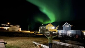Une personne se tient dans une cour, avec des maisons en arrière-plan et des aurores boréales dans le ciel.