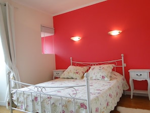 Un dormitorio con estructura de cama metálica, ropa de cama floral, dos mesitas de noche y una pared roja.