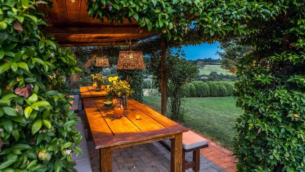 Mesa de madera al aire libre con un florero y una jarra, bajo una pérgola de madera con cestas colgantes, rodeada de abundante follaje y vistas a un campo cubierto de hierba.