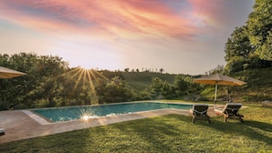 Una piscina con rayos de sol, rodeada de césped y árboles, y dos sillas de jardín con sombrillas.