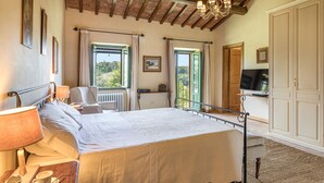 Un dormitorio con una cama grande, una silla, un escritorio, un televisor y una ventana con cortinas.