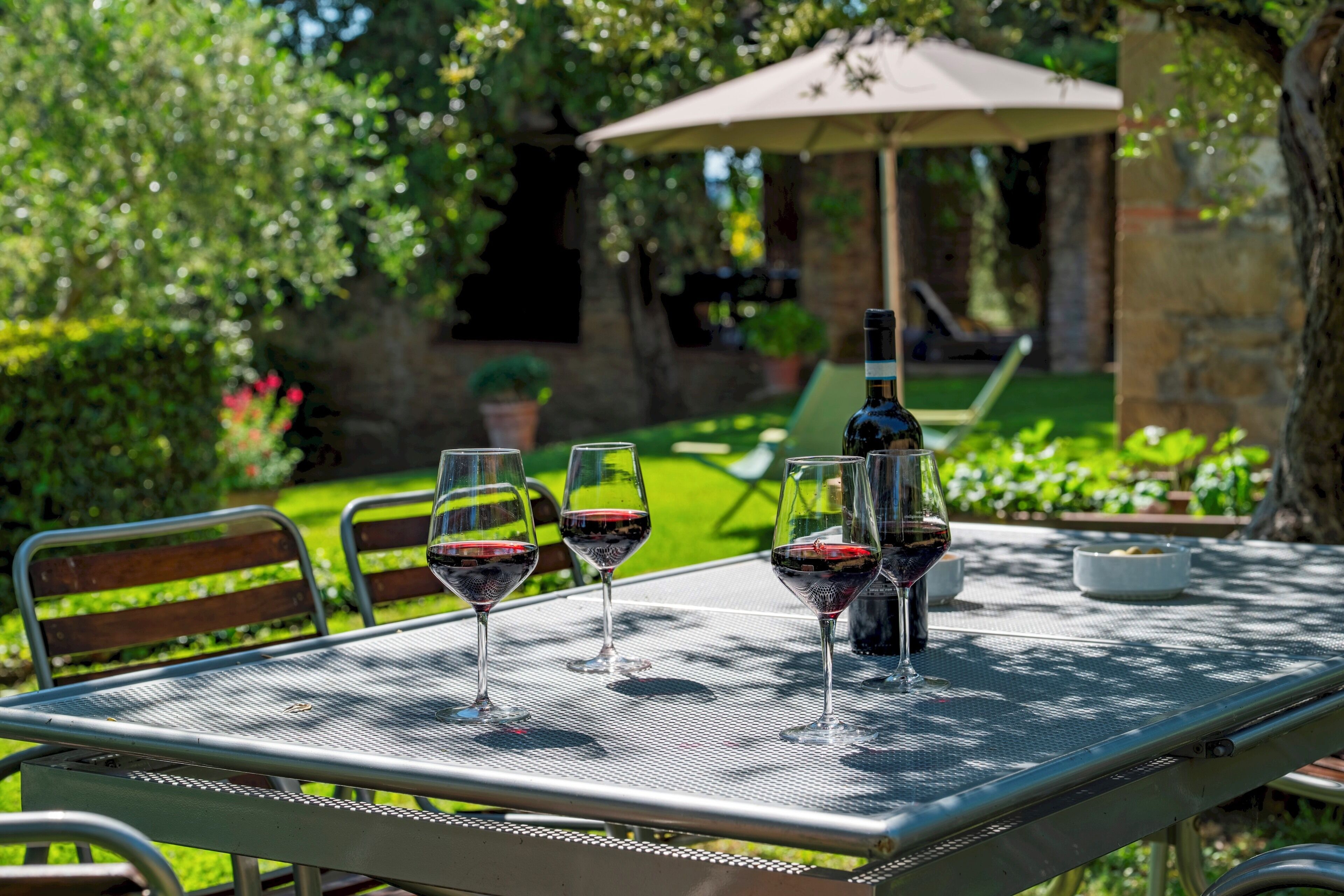 A metal table with three wine glasses and a bottle, set outdoors with a green lawn and a stone wall in the background.