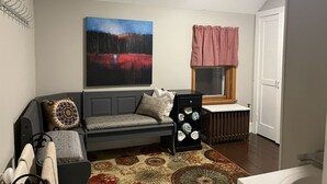 Un salon moderne avec un canapé gris, des coussins à motifs, un grand tableau abstrait, une fenêtre agrémentée d’une lambrequin rouge et un radiateur.