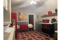 Une cuisine compacte avec un réfrigérateur rouge, un four à micro-ondes et une machine à café placés sur une étagère. Un tapis à motifs recouvre le sol, et une porte donne accès à une autre pièce.