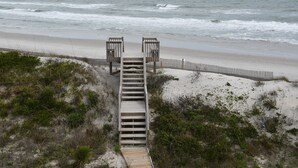 Un escalier en bois mène à une plage, qui est bordée d’une clôture en bois et surmontée de deux constructions en bois.