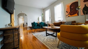 A modern living room with a wooden floor, teal and yellow chairs, a round coffee table, and artwork on the walls. - Cozy 3Bed Home w/ Firepit By Zoo, Downtown & I/80❤ (Omaha)