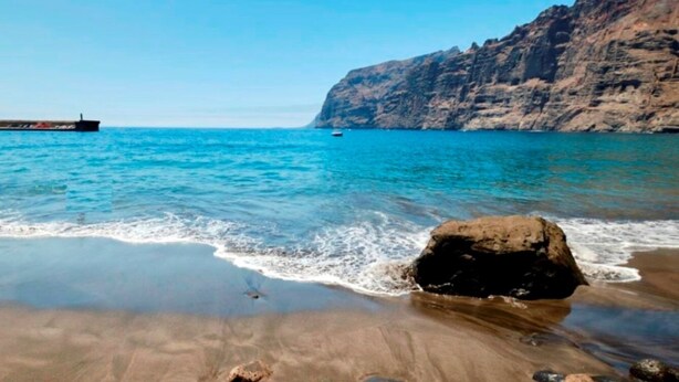 A beach with clear blue water, a large rock in the foreground, and a cliff in the background.
