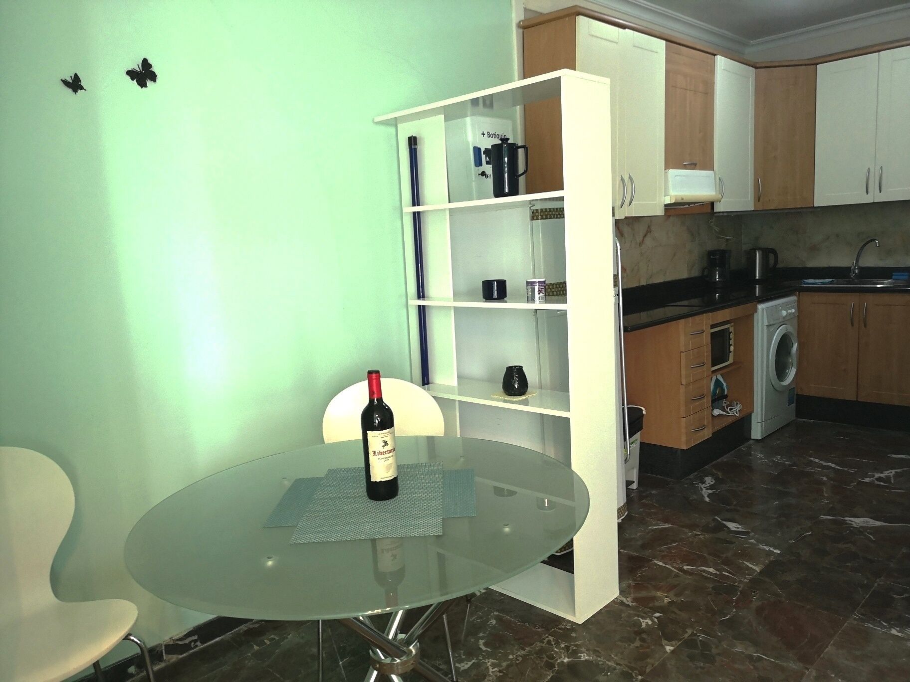 Una cocina pequeña con lavadora, cafetera y una botella de vino sobre una mesa de vidrio.