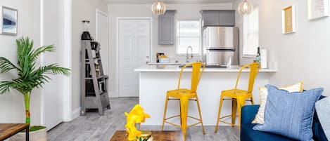 Eine moderne Küche mit weißen Schränken, einem Edelstahlkühlschrank und einem eingebauten Backofen. Im Wohnbereich befinden sich zwei gelbe Stühle und ein blaues Sofa.