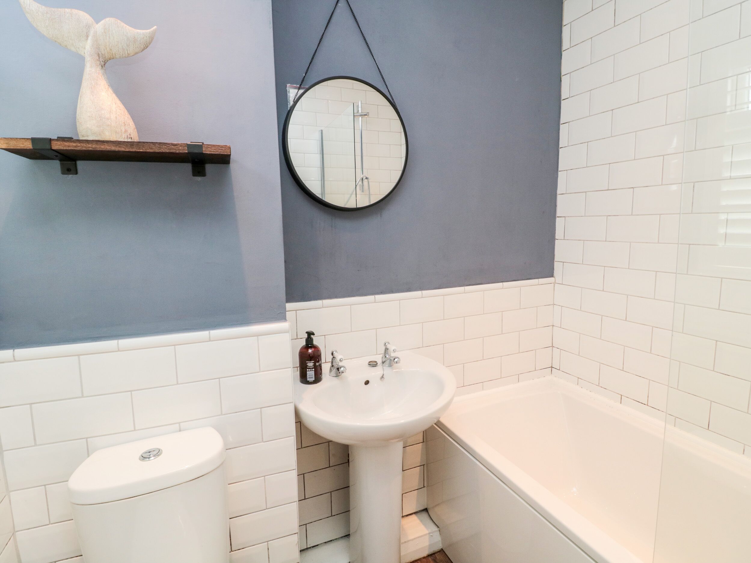 A bathroom with a toilet, sink, and bathtub. There is a wooden shelf with a decorative item above the sink.