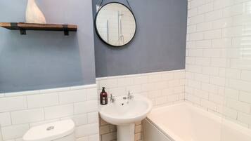 A bathroom with a toilet, sink, and bathtub. There is a wooden shelf with a decorative item above the sink.