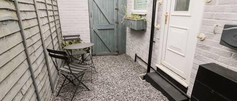 A small outdoor seating area with a table and chairs, a wooden fence, a door, and a gravel floor.