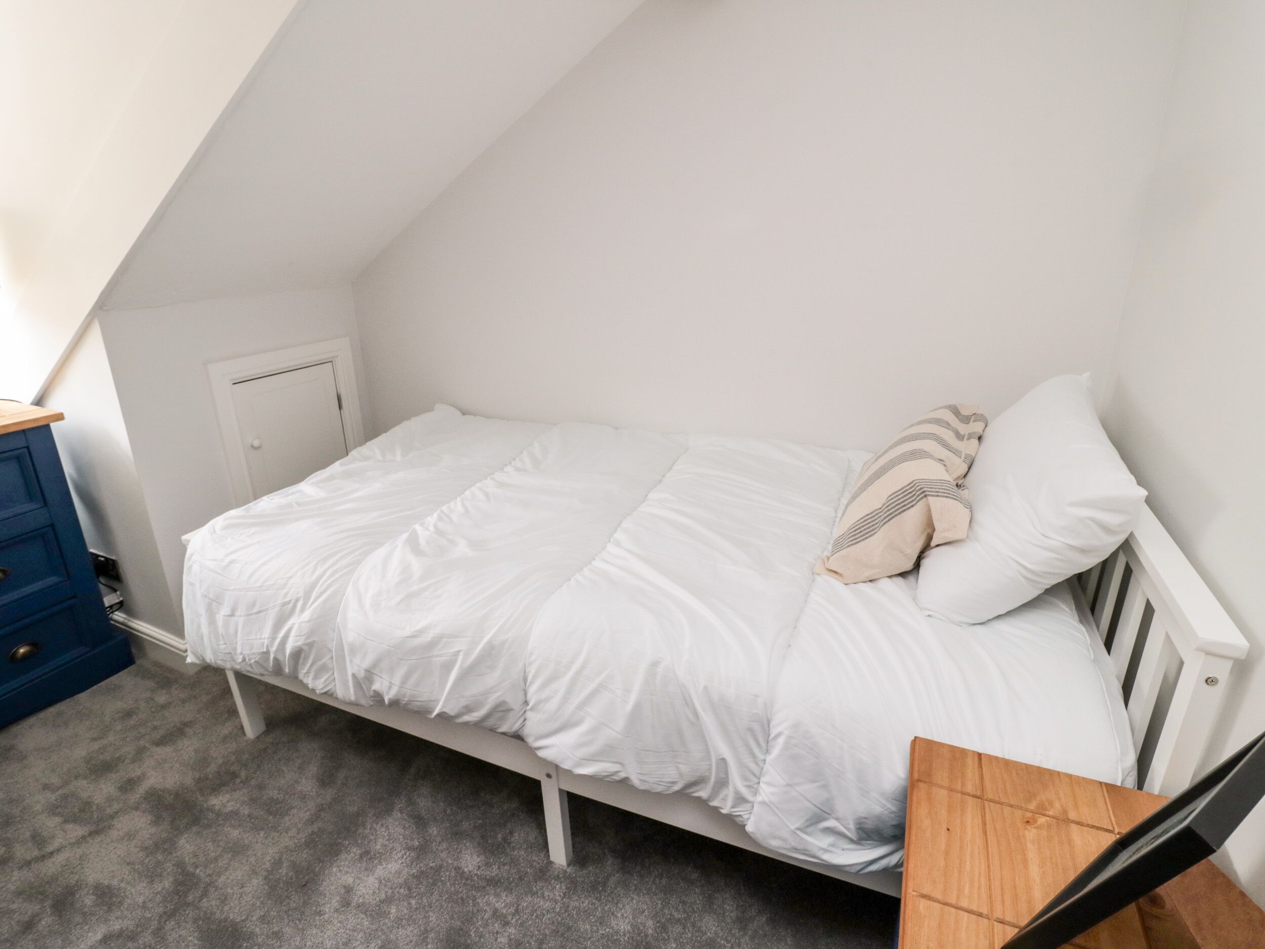 A small, white, single-bed room with a sloped ceiling, a blue dresser, and a small table.