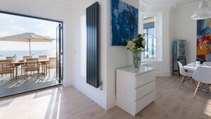 Un salon moderne doté d’une grande porte-fenêtre coulissante donnant sur une terrasse extérieure, d’une console blanche ornée d’un vase de fleurs et d’un grand tableau abstrait accroché au mur.