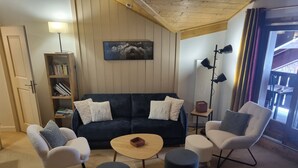 Un salon chaleureux avec un canapé bleu foncé, deux fauteuils blancs, une table basse ronde et un petit pouf. Une bibliothèque et un tableau encadré ornent le mur.
