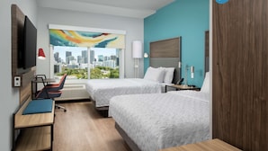 Habitación de hotel con dos camas, un escritorio, una silla, un televisor y ventana con vista a la ciudad.