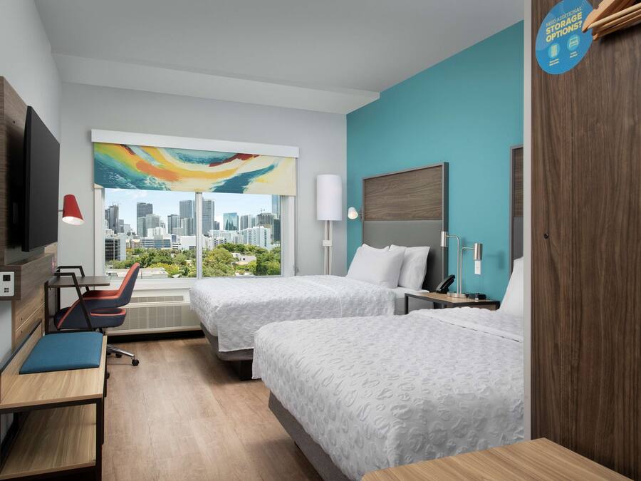 A hotel room with two beds, a desk, a chair, a TV, and a window with a city view.