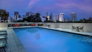 Una piscina en la azotea con sillones y mesas, rodeada por el horizonte de una ciudad al atardecer.