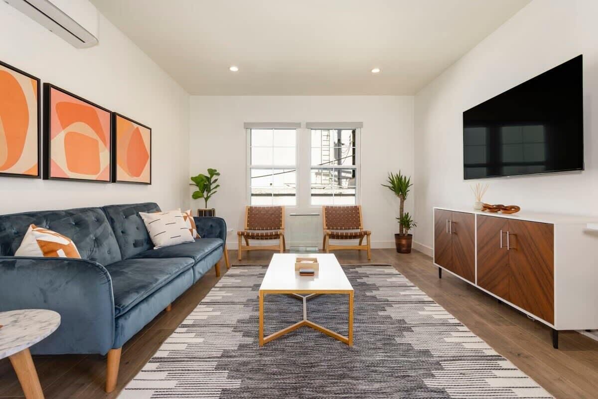 Una sala moderna con un sofá azul oscuro, una mesa de centro blanca y un televisor montado en la pared. Hay dos sillones y un mueble con televisor.