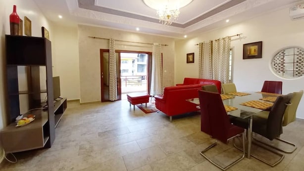A spacious living room with a red sofa, a dining table with chairs, a TV on a stand, and a chandelier. - Lovely 2-bed Apartment in Kribi-douala (Kribi)
