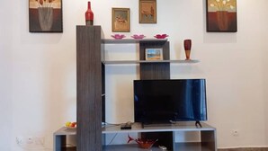 A modern living room with a flat-screen TV on a wooden stand, surrounded by shelves with decorative items and framed pictures. - Lovely 2-bed Apartment in Kribi-douala (Kribi)