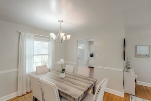 A dining area with a wooden table, white upholstered chairs, a chandelier, and a view into the kitchen.