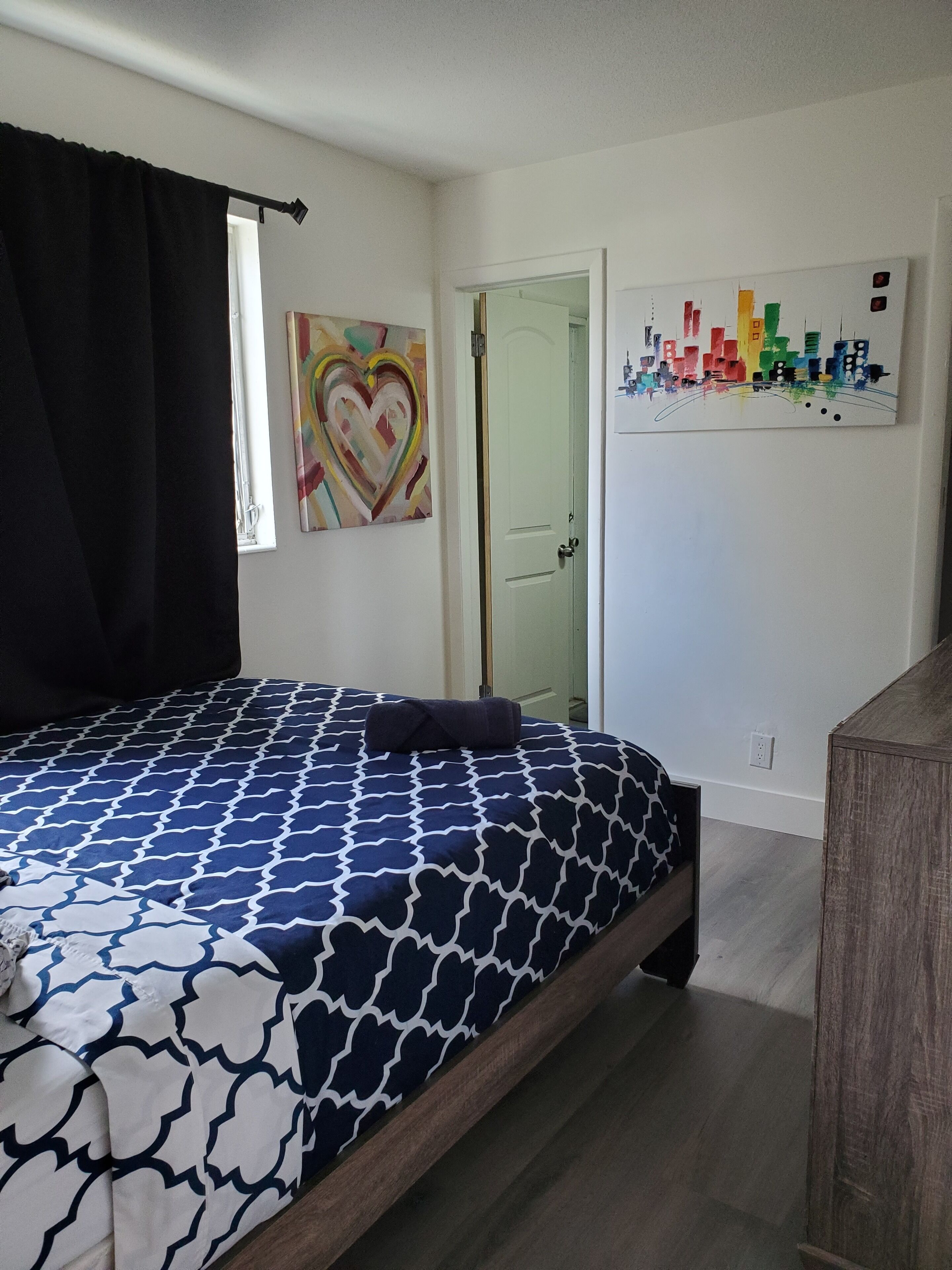 A bedroom with a bed, a door, and a wall with a heart-shaped painting and a cityscape painting.