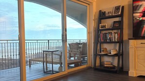 Een woonkamer met een grote schuifpui die uitkomt op een balkon met tuinmeubilair. Er is een boekenkast met boeken en decoratieve objecten, en een televisie aan de rechterkant.